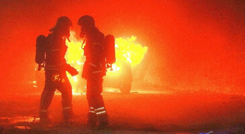Zwei Männer in Feuerwehrausrüstung befinden sich untertage vor einem brennenden Fahrzeug.